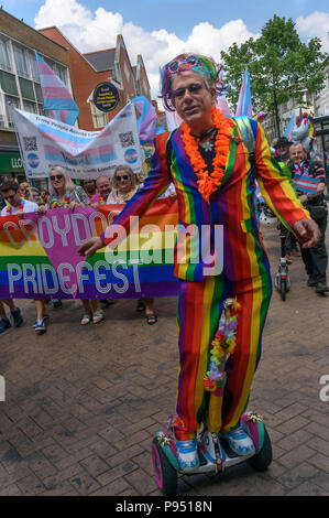 London, Großbritannien. 14. Juli 2018. Ein Mann in einem Regenbogen Anzug reitet ein HOVERBOARD an der Vorderseite des Croydon Stolz Prozession. Mehrere hundert Menschen zogen durch die Innenstadt von Croydon auf dem Weg in das dritte Croydon Croydon Pridefest, durch Rat gefördert, in Wandle Park. Viele wurden in Buntes Kleid und es gab Banner, Fahnen, Schilder, Plakate und Einhörner. Das kostenlose Festival sollen LGBT + Gleichheit und Vielfalt in Croydon, zu fördern, und ist Londons zweitgrößte Pride Festival. Nach der Unterbrechung durch Anti-Trans-Aktivisten in London Pride, der Parade gab den Trans Menschen in London (TRA Stockfoto