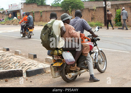 Kamdini, Uganda - Straßenszene mit Menschen und Motorräder. Drei Männer fahren mit dem Motorrad. Stockfoto