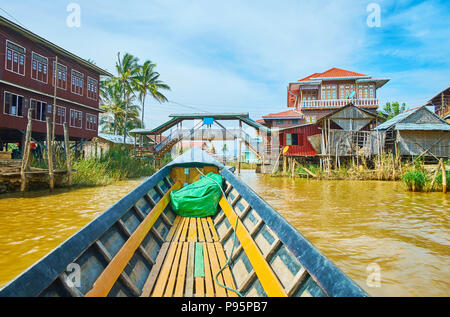 Kanu schwimmt entlang der hölzernen Cottages von Ywama Dorf auf dem Inle See mit Blick auf die Brücke auf dem Hintergrund, Myanmar. Stockfoto