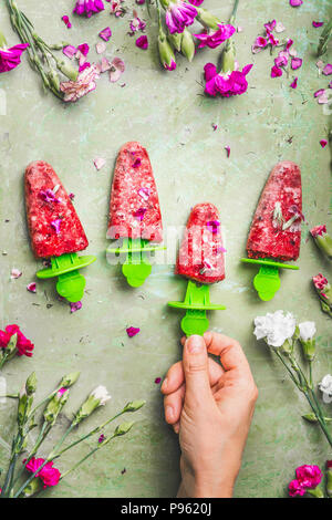 Weibliche Hand, die hausgemachte Obst und Beeren Eis oder Eis am Stiel auf Teal rustikalen grünen Tisch Hintergrund, mit Garten Blumen, Ansicht von oben eingerichtet. Stockfoto
