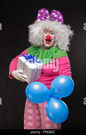 Lustige Clown mit zotteligen Haaren und einem fröhlichen Make-up hält ein Geschenk auf schwarzem Hintergrund Stockfoto