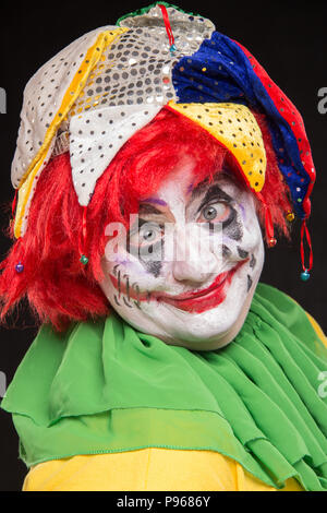 Ein schrecklicher Clown mit einem furchtbaren Make-up und Hut auf dem Kopf lachend auf schwarzem Hintergrund Stockfoto