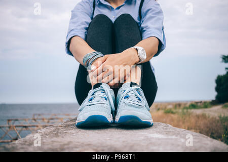 Junge Frau sitzt auf der Brüstung in der Nähe vom Meer umarmt die Knie. Weibliche hipster tragen blaue T-Shirt, Turnschuhe und eine schwarze Hose mit einem Rucksack Kreuzung h Stockfoto