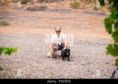 Schönen weißen Haare ältere Frau kaukasischen Sitzen mit zwei nette lustige Mops Vielfalt Rassen und Farben in Outdoor Freizeitaktivitäten Aktivität im Sommer Stockfoto