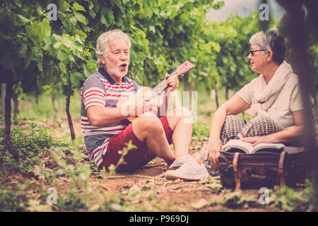 Paar Alternativen im Alter von älteren Reisenden bleiben oder sich in einen Weinberg mit dem Gepäck und spielen eine Ukulele, E-Gitarre und Lesen ein Papier Stockfoto