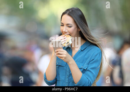 Glückliche Frau essen einen Burger mit einem Smart Phone auf der Straße Stockfoto