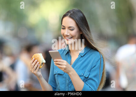 Frau mit einem Burger Kontrolle smart phone auf der Straße Stockfoto