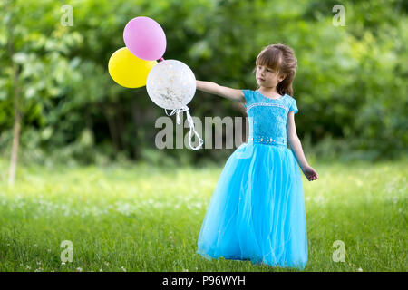 Schöne kleine Blonde langhaarige Mädchen in schönen langen blauen Abendkleid suchen wie Prinzessin schaut in die Kamera halten bunte Luftballons in Bl Stockfoto