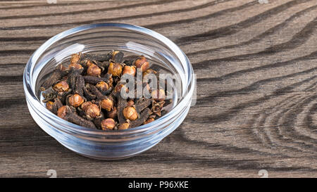 Close-up würzig aromatische Gewürznelken in Glasschale. Syzygium aromaticum. Stapel von getrockneten Heilpflanzen Gewürze mit markanten Duft auf braunem Holz Hintergrund. Stockfoto