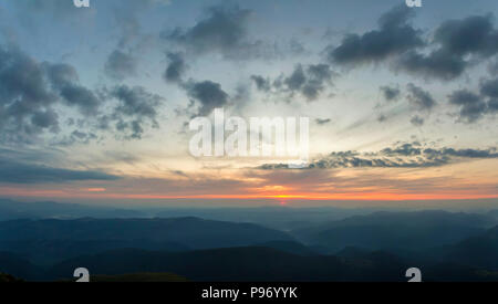 Fantastischer Sonnenaufgang oder Sonnenuntergang über grünen Bergrücken mit dichten blauen Nebel bedeckt. Helles orange sun Erhöhung in weichem bewölkter Himmel über Horizont. Stockfoto