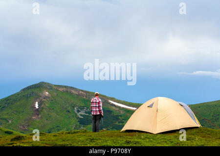 Jungen gutaussehenden Mann stehen auf der grünen Wiese Tal an einem sonnigen Tag an der kleinen Hütte in den Bergen und der strahlend blaue Himmel Hintergrund. Tourismus, Reisen, Hi Stockfoto