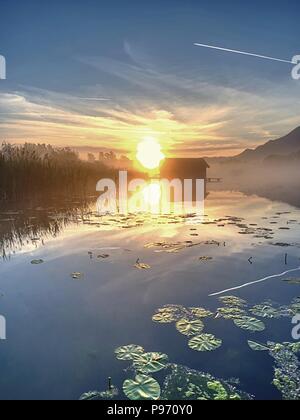 Romantischen sonnenaufgang Reflexion auf dem Kochelsee gegen Bootshäusern. Anfang Sommer morgen ansehen, Bayern, Deutschland Stockfoto