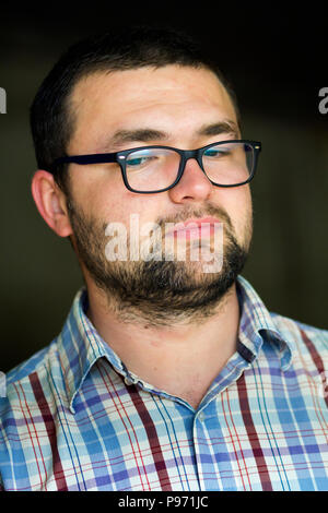 Portrait von gut aussehenden bärtigen Schwarzhaarige intelligente moderne junge Mann in Gläsern mit kurzen Haarschnitt und eine Art schwarze Augen lächelnd in die Kamera auf verschwommen Stockfoto