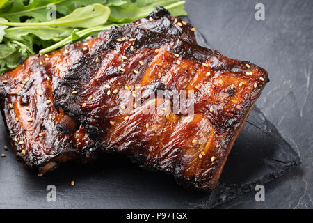 Nahaufnahme von Rippchen vom Grill mit BBQ Sauce und Rucola. Leckeren Snack zum Bier auf einem Stein Vorstand für die Einreichung auf dunklen konkreten Hintergrund Stockfoto