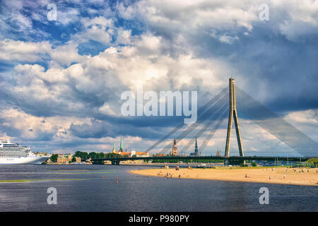 Sandstrand am Ufer des Flusses Daugava auf dem Hintergrund der Schrägseilbrücke und das Panorama der Stadt mit Turmspitzen und Schiff im Hafen. Stockfoto