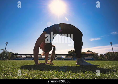 Eine junge Frau übernimmt die Gymnastik Yoga Übungen in das städtische Stadion. Athlet Turnerin stand auf der Brücke auf dem Gras Stockfoto