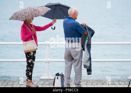 Aberystwyth Wales UK, Montag, 16. Juli 2018 UK Wetter: Nach vielen Wochen der fast ständigen Sonnenschein und außergewöhnlich trockenes Wetter, der Regen mit Leuten gesehen, die Zuflucht vom Regenguß unter ihren Schirmen, wie Sie entlang der Promenade in Aberystwyth auf der West Wales Küste Foto credit Spaziergang zurückgekehrt: Keith Morris Alamy leben Nachrichten Stockfoto