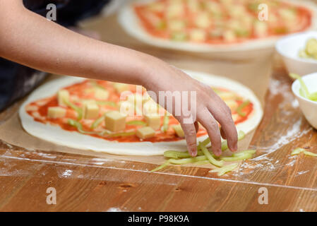 In der Nähe von Kinderhänden Vorbereitung Pizza. Kinder legen Sie auf der Grundlage der Pizza Gemüse und Käse Stockfoto