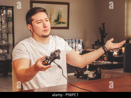 Gamer Mann hält eine Steuerung mit einem empörten Ausdruck wie es Fragen einige Ergebnis auf dem Spiel. Mann spielen Im video Spiel zu Hause. Stockfoto