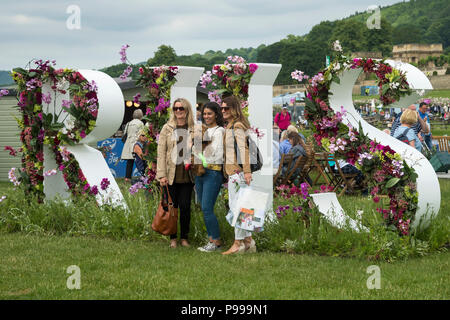 3 Frauen & Hund, lächelnd und für Fotos posiert, stehen vor großen Blumenarrangement (Buchstaben R H S) - Chatsworth Flower Show, Derbyshire, England, UK. Stockfoto