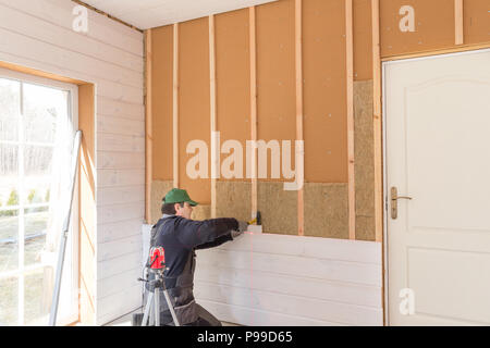 Der Arbeitnehmer macht Ausbauarbeiten der Wände mit einem weißen Holzplatte mit Laser Line Level. Gebäude wärmedämmenden eco-Rahmen aus Holz haus mit Holzfaser Platten Stockfoto