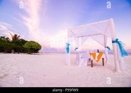Romantisches Abendessen Tisch Set-up für eine Hochzeitsreise Paar am Strand unter Sonnenuntergang Himmel. Exotische Hochzeit und Flitterwochen Konzept Stockfoto