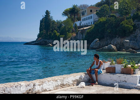 Junge sitzt auf dem Bootssteg in dem hübschen kleinen Hafen Dorf Loggos, Paxos, Griechenland Stockfoto