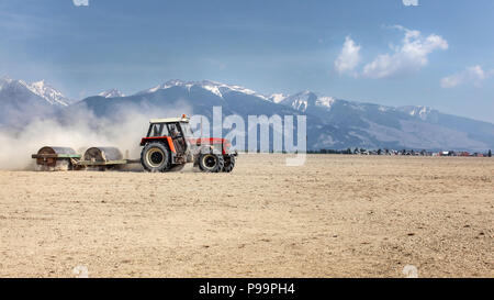 Tractor Pulling heavy metal Walze über trockenes Feld an einem schönen Frühlingstag mit Bergen im Hintergrund. Die Bodenbearbeitung. Stockfoto
