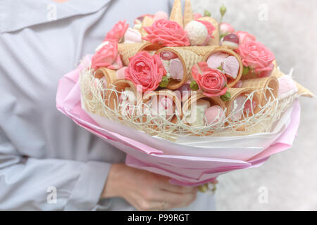 Mädchen hält in der Hand einen schönen Blumenstrauß, bestehend aus rosa Blüten einer Rose und verschiedenen Süßigkeiten als Geschenk Stockfoto