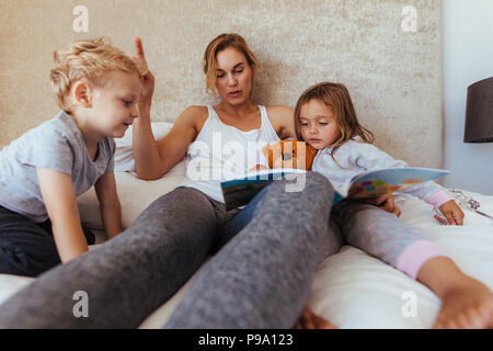 Mutter Lesung Gute-Nacht-Geschichte für Kinder zu Hause. Fürsorgliche Mutter lesen Buch für entzückende kleine Kinder auf dem Bett. Stockfoto