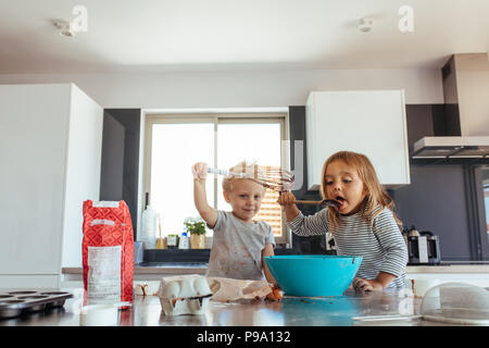 Junge Mädchen lecken Teig von Spachtel mit Ihrem Bruder mit einem Schneebesen. Geschwister, Kuchenteig in der Küche. Stockfoto