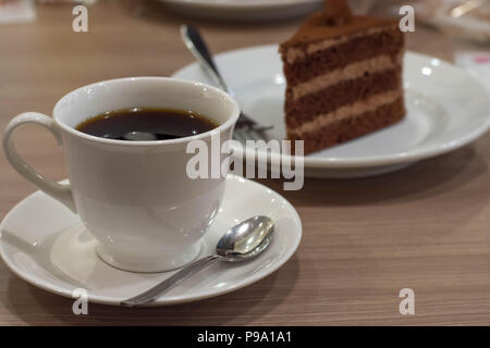 Eine Tasse schwarzen Kaffee auf einer Untertasse mit mini Löffel und ein Stück Schokoladenkuchen auf weiße Platte im Hintergrund Stockfoto