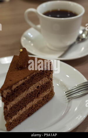 Schokoladenkuchen mit essbaren Gold auf weißer Teller mit Gabel und Tasse Kaffee im Hintergrund auf einem Tisch. Stockfoto