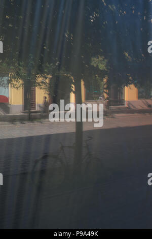 Reflexion der ein Fahrrad in der Nähe von einem Baum auf der Straße mit Schatten in einen Film mit Falten im Glas ein Schaufenster Stockfoto