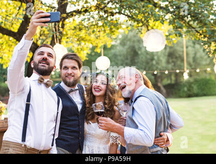 Braut, Bräutigam und Gäste mit Smartphone unter selfie außerhalb an der Hochzeit. Stockfoto