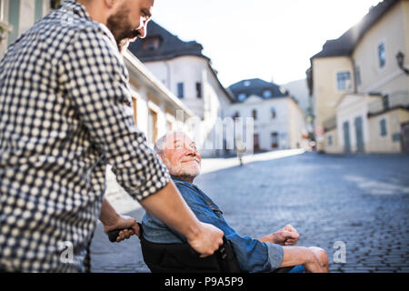 Ein erwachsener Sohn mit dem Vater im Rollstuhl auf einen Spaziergang in der Stadt. Stockfoto