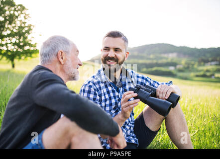 Ein erwachsener Sohn Hipster mit Fernglas und Senior Vater sitzen auf dem Gras in sonnige Natur. Stockfoto