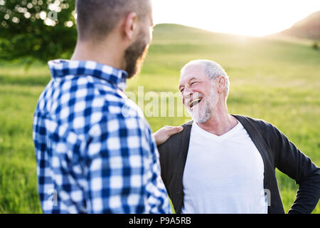 Ein erwachsener Sohn Hipster mit Senior Vater auf einem Spaziergang in der Natur bei Sonnenuntergang. Stockfoto