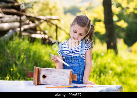 Ein kleines Mädchen mit einem Pinsel außerhalb, malen eine hölzerne birdhouse. Stockfoto