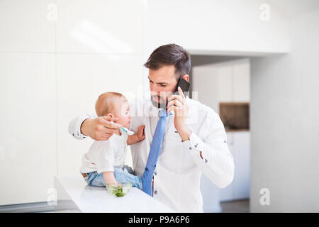 Vater mit Hemd und Krawatte und Smartphone Fütterung ein Baby Sohn zu Hause, wenn Sie das Gespräch wieder auf. Stockfoto