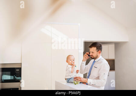 Vater mit Hemd und Krawatte und Smartphone Fütterung ein Baby Sohn zu Hause, wenn Sie das Gespräch wieder auf. Stockfoto