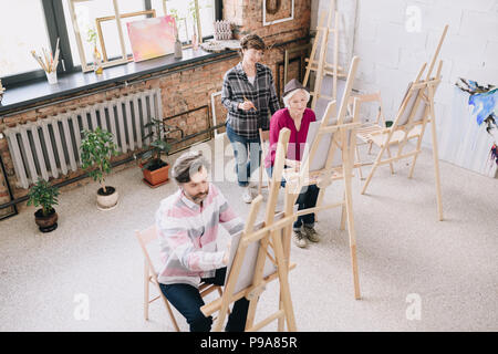 Hohen winkel Porträt von zwei erwachsenen Kursteilnehmer Malerei sitzt von staffeleien in art studio mit weiblichen kunst Lehrer beobachten Sie in geräumigen und lichtdurchfluteten Loft spac Stockfoto