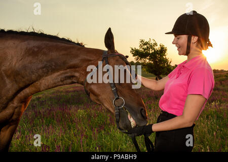 Schöne lächelnde Mädchen Jockey stand neben ihr braunes Pferd Tragen besonderer Uniform auf einen Himmel und grünen Feld Hintergrund auf einen Sonnenuntergang. Stockfoto