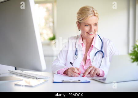 Nahaufnahme der lächelnden älteren Ärztin mit Ihrem Laptop und schreiben etwas, während am Schreibtisch beim Arzt im Wartezimmer sitzen. Stockfoto