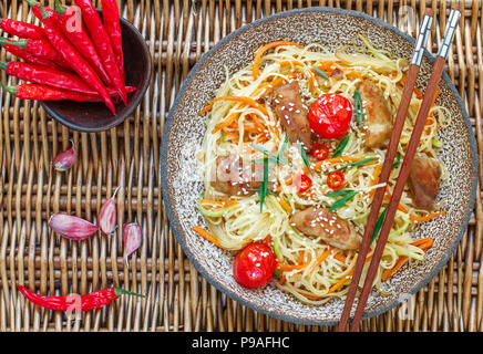 Rühren gebratene Nudeln mit Huhn, Gemüse (Karotten, Tomaten, Zucchini, Knoblauch, Paprika, grüne Zwiebeln), Sesam und Soja Sauce auf Lehm Platte. Asiatische Stockfoto