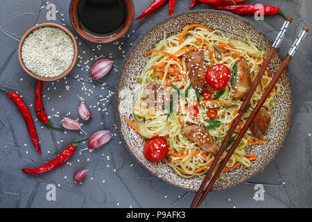 Rühren gebratene Nudeln mit Huhn, Gemüse (Karotten, Tomaten, Zucchini, Knoblauch, Paprika, grüne Zwiebeln), Sesam und Soja Sauce auf Lehm Platte. Asiatische Stockfoto