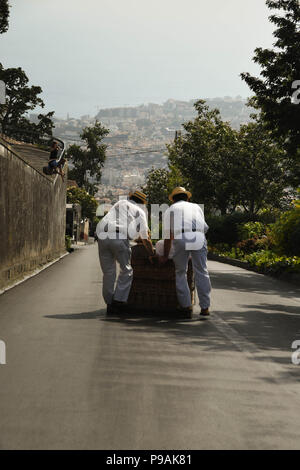 Touristen genießen die Fahrt bergab auf einem traditionellen Weidenkorb Rodeln auf den Straßen über Funchal, Madeira Stockfoto