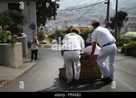 Touristen genießen die Fahrt bergab auf einem traditionellen Weidenkorb Rodeln auf den Straßen über Funchal, Madeira Stockfoto