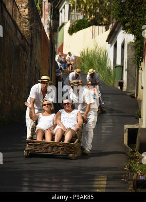 Touristen genießen die Fahrt bergab auf einem traditionellen Weidenkorb Rodeln auf den Straßen über Funchal, Madeira Stockfoto