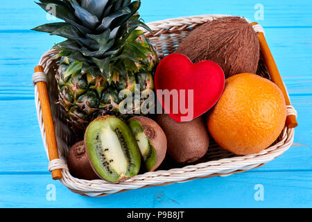 Zusammensetzung aus tropischen Früchten und roten Herzen. Sortierte Früchte im Korb auf blauen Holzmöbeln. Früchte Erfrischung und Sommer. Stockfoto